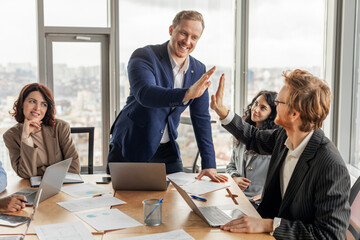 Aufkleber - A businessman in a blue suit gives a high five to a colleague in a black suit during a meeting in a modern office. Other colleagues are present and working at a table with laptops and papers