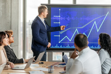 Aufkleber - A businessman in a suit points to an upward trending line graph on a large screen while presenting to his team during a meeting. His team sits around a table with laptops open
