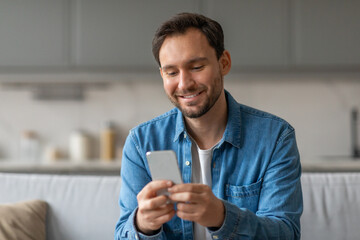 Poster - A man wearing a blue shirt is sitting on a couch in a living room, smiling while using his smartphone.