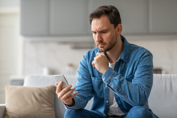 Poster - A man sits on a couch in a living room, looking concerned as he reads his smartphone. He is wearing a blue shirt and jeans. His hand is on his chin and his brow is furrowed.
