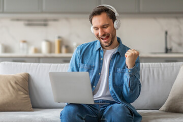 Poster - A man wearing headphones and a blue shirt sits on a couch in a living room and celebrates a win on his laptop. He smiles with excitement while raising his fist in the air.