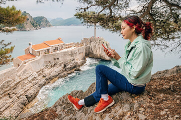 Wall Mural - Woman using a smartphone map application at a seaside viewpoint in historic Petrovac, Montenegro