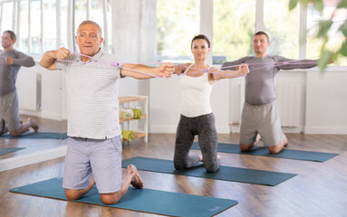 Wall Mural - Elderly man doing pilates exercises with elastic loop in group in fitness studio