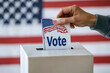 © Phichitpon - man is voting in an USA election by putting a white piece of paper into a box. USA flag on background.