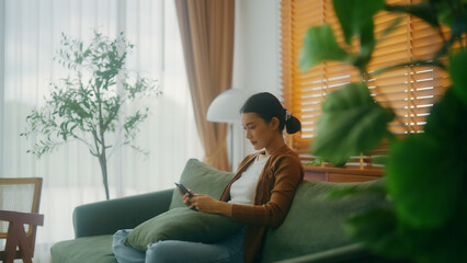 Smiling pretty young Asian woman sitting on cozy couch, using funny mobile apps in living room. Woman at home, Doing online shopping, Messaging friends, Posting on social Media