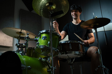 A man is sitting behind a drum set, playing the drums. He is wearing a black t-shirt and shorts. The drum set is green, and the room appears to be a studio, possibly soundproofed