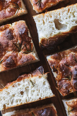 Detailed close-up of sliced focaccia bread pieces on a wooden surface, showcasing the bread's airy texture and crisp, golden-brown crust. Sprigs of rosemary are visible on the surface.