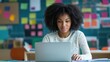 © keetazalay - A young African American teacher sitting at her desk in a modern classroom, working on a laptop, preparing lesson plans, and utilizing digital resources to enhance her teaching methods
