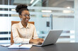 © Liubomir - Young African American businesswoman wearing glasses working on laptop at modern office, smiling, looking motivated, professional, and engaged. Concept workplace productivity, professional success.
