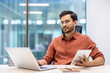 © Liubomir - Serious businessman working with laptop and tablet in modern office. Focused on tasks, using both devices to enhance productivity. Professional setting with large windows and natural light.