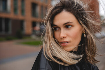 Close up portrait blonde short hair cut woman with blowing on wind hair walking outdoor. Sensual and dreaming girl look thoughtful. She wear black leather shirt.