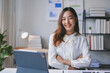 © amnaj - Young asian businesswoman smiling while sitting at her desk with her arms crossed in a modern office
