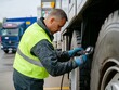 © Nathan - Male worker in a reflective vest checks truck tire pressure at a service station.