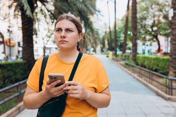 Wall Mural - Negative people emotion. Upset young 30s woman is using a smartphone outdoors. Woman tourist dressed in casual look holding smartphone gadget in hand for communicate on the city street.