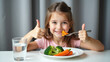 © Nikolaus - A cheerful young girl with a bright smile gives a thumbs up while eating a plate of healthy vegetables, including carrots and broccoli, promoting nutritious eating.