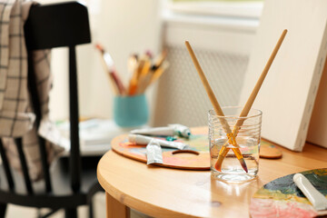 Poster - Artist's palette, brushes in glass of water and paints on wooden table indoors. Space for text