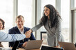 © Prostock-studio - A businesswoman in a grey blazer leans across a conference table to shake the hand of a male colleague while another colleague sits across from them. All three people are smiling