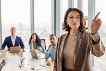 Aufkleber - A confident businesswoman wearing a brown blazer stands and gestures with her right hand while giving a presentation during a meeting. She is surrounded by colleagues seated at a table