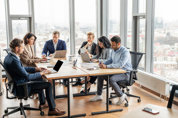 Aufkleber - A group of business professionals work on their laptops at a large wooden table in a modern office. The office is filled with natural light from large windows overlooking a city.