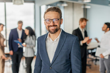 Aufkleber - A businessman wearing a blue suit and glasses smiles while standing in a modern office setting. Other office workers are standing in the background and engaged in conversations.