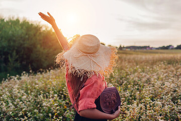 Wall Mural - Back view of happy stylish woman wearing straw hat in blooming buckweat field at sunset holding leather handbag.