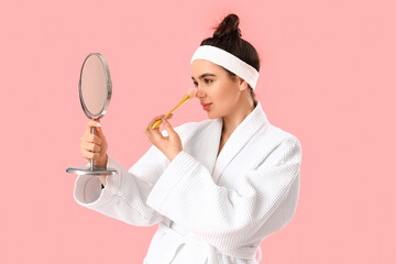 Poster - Pretty young woman in bathrobe applying makeup on pink background