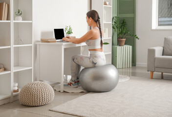 Poster - Sporty young Asian woman using laptop while sitting on fitball at home