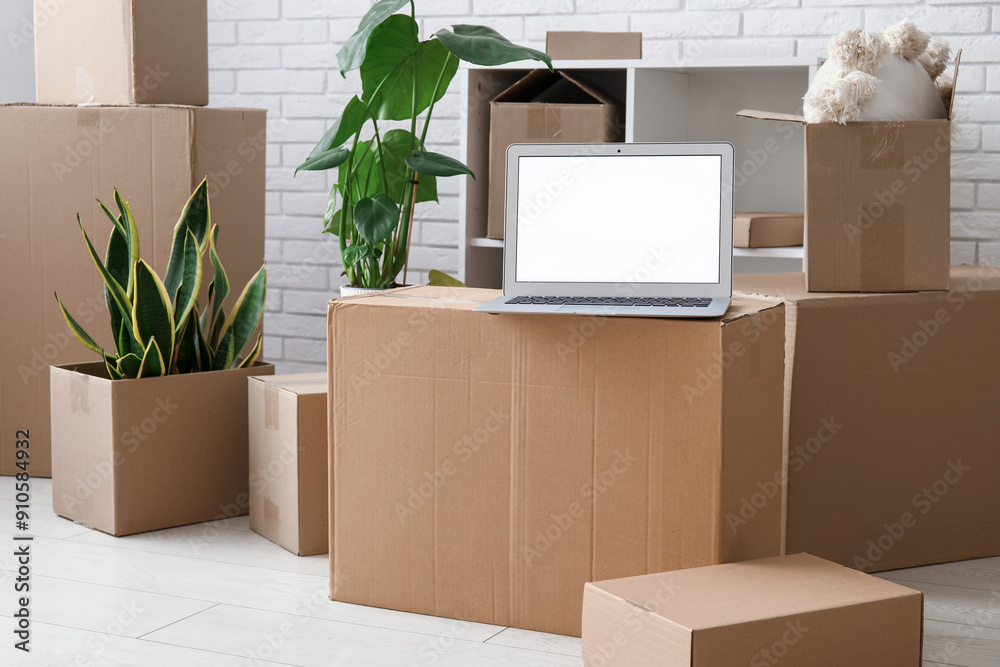 Blank laptop with plants and moving boxes near white brick wall in room
