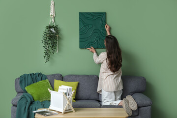 Wall Mural - Young pretty woman hanging picture on green wall at home