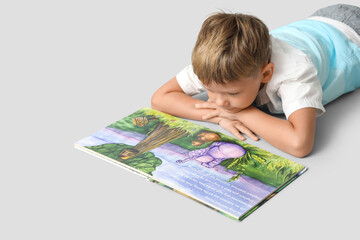 Wall Mural - Portrait of cute little boy lying on floor and reading book on grey background