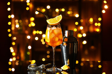 Wall Mural - Glass of Aperol spritz cocktail and shaker on table against blurred lights