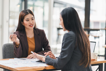 Wall Mural - Two businesswomen in formal attire having discussion at desk with charts and graphs, focusing on data analysis and teamwork.