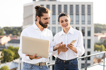 Wall Mural - Young professionals discussing work while using laptop and smartphone in outdoor urban setting. Showing teamwork, collaboration, and tech-savvy approach to business. Cityscape in background.