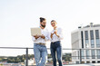 © sofiko14 - Young man and woman standing outdoors discussing work on laptop and smartphone. Casual attire, urban setting, teamwork and collaboration. Modern business concept.