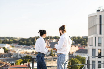 Wall Mural - Business colleagues standing on rooftop, discussing project using technology devices. Urban cityscape view in background provides professional setting. Teamwork and collaboration concepts represented.