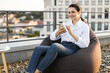 © sofiko14 - Woman sitting comfortably on bean bag on rooftop, smiling while using smartphone. Casual and relaxed setting with view of buildings in background, showcasing leisure and technology use.