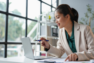 Happy Asian businesswoman in office working on financial documents, holding pen pointing at laptop. Results, income, online marketing. Company taxes Online business work.