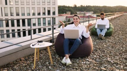 Wall Mural - Two professionals working on laptops seated on bean bags on rooftop. Relaxed and productive outdoor workspace with cityscape view. Concept of remote work team collaboration and modern work environment