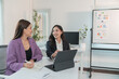 © Tj - Two Asian businesswomen cheerfully discussing plans in office using calculator and laptop, standing in front of a whiteboard talking.
