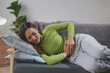 © amnaj - Young woman lies on the sofa, holding her stomach in pain at home, looking unhappy and worried about a medical issue