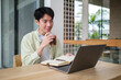 © wattana - A young man is seated at a wooden table, working on his laptop with a notebook open beside him.