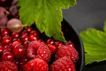 Tasty fresh ripe raspberry, red currant, gooseberry, green leaves. Nutrients, Healthy berry texture on dark background. Red summer berries in black bowl, high quality photo