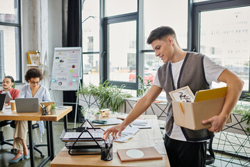 Wall Mural - Young elegant man packing his items during lay off, colleagues on backdrop.