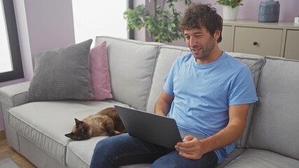 Wall Mural - A cheerful hispanic man works on a laptop beside a resting siamese cat on a living room couch at home.