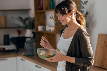 Pretty smiling woman eating healthy salad standing in the kitchen.