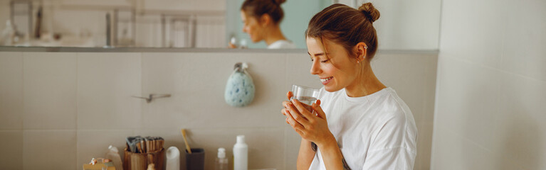 Wall Mural - Young beautiful woman drinking glass of water in the bathroom at home. Beauty treatment