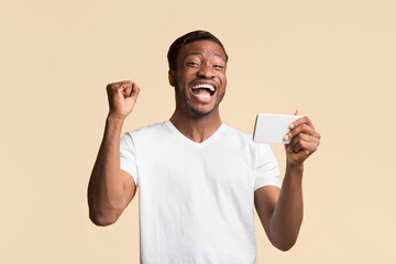 Poster - Excited Afro Man Holding Smartphone Shaking Fists Winning Mobile Game Standing Over Yellow Studio Background.