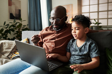 Wall Mural - Young African American man sitting on sofa next to his little son while both looking at laptop screen and watching online video at home