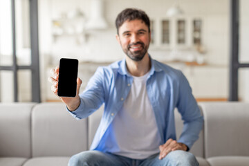 Poster - A man wearing a blue shirt is seated on a gray couch in a living room. He is smiling and looking directly at the camera. He is holding his smartphone in front of him