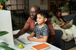 © pressmaster - Young African American man in casual attire embracing his cute little son while both sitting by desk in front of computer monitor